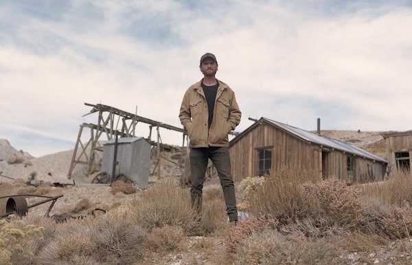 Man standing confidently in front of an old mining structure in a ghost town, wearing a jacket and cap.