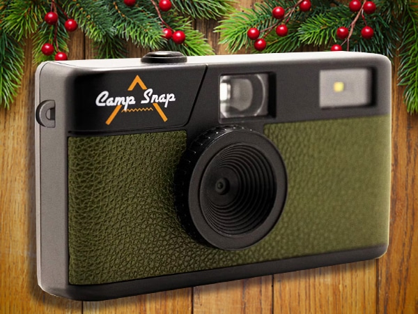 A green and black Camp Snap camera is displayed in front of a wooden background decorated with pine branches and red berries—an ideal choice for stocking stuffers for men this holiday season.