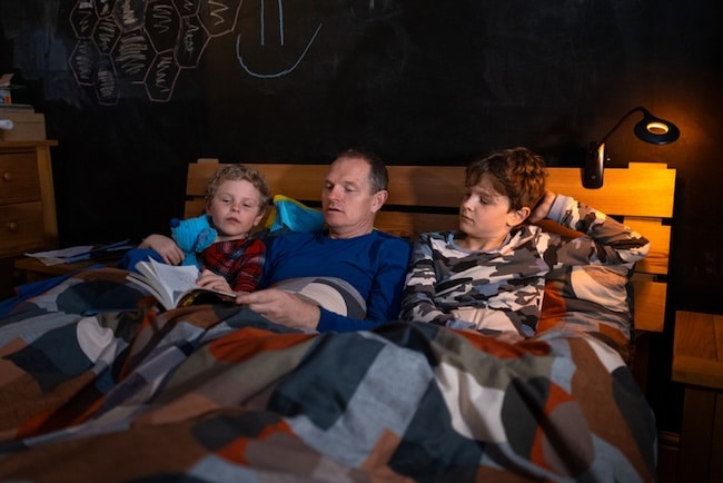 An adult and two kids sit in bed under a patterned duvet, reading aloud from classic chapter books, with a wall light and chalk drawings visible in the background.