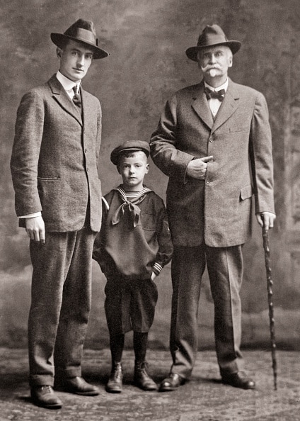 Three males from different generations pose for a formal portrait, showcasing timeless men's style; a young boy in a sailor suit stands between two men in suits and hats, one older with a cane.