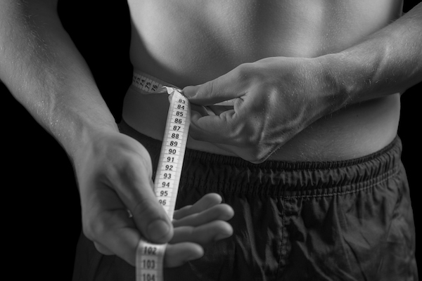 A man is measuring his waist with a measuring tape to track his body fat.