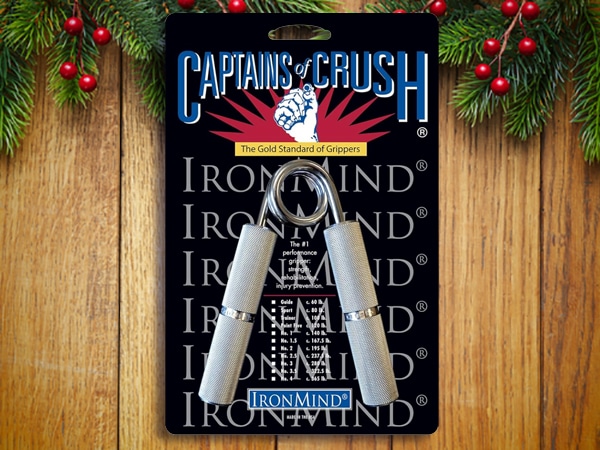 A packaged Captains of Crush hand gripper by IronMind is displayed on a wooden surface with evergreen branches and red berries, making it one of the perfect stocking stuffers for men this holiday season.