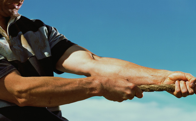 Close-up of two people engaged in a tug of war, highlighting their arms and the rope as they develop grit.