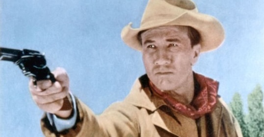 A man in a cowboy hat and bandana, embodying the spirit of the American gunfighter, points a revolver with a serious expression, outdoors under a clear blue sky.