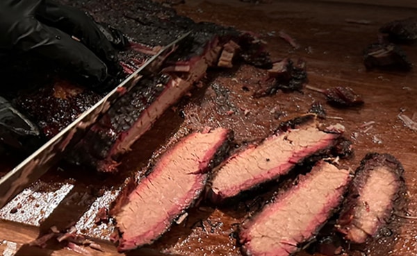 Sliced smoked brisket rests on a wooden cutting board, with a gloved hand deftly holding a knife—perfect for your weekend BBQ.