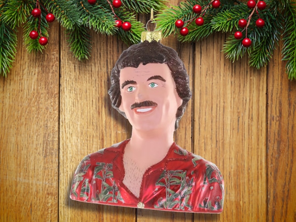 A Christmas tree ornament shaped like a man's bust with curly hair, a mustache, and a red patterned shirt—perfect as unique stocking stuffers for men—hangs in front of a wooden background with pine branches and red berries.