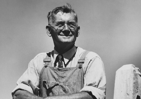 Black and white photo of an older man wearing glasses, a tie, and overalls. He is smiling with arms crossed in front of him against a clear 1930s sky.