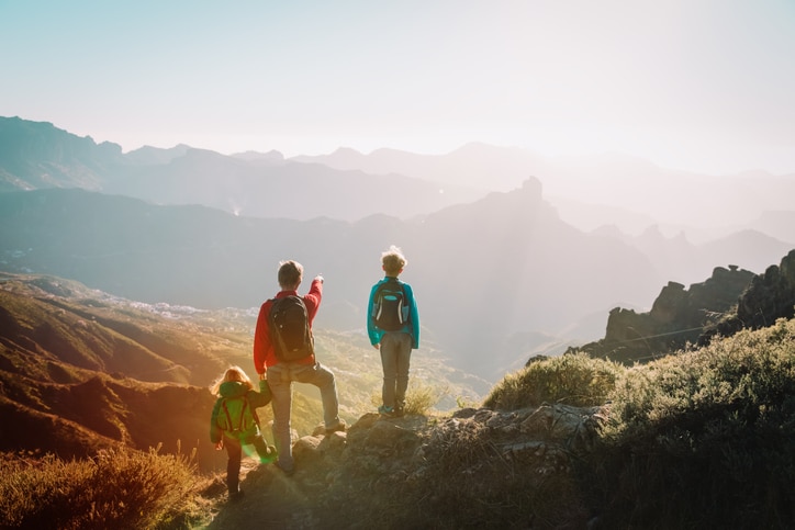 Three people with backpacks stand on a mountain trail, overlooking a scenic valley bathed in sunlight. One, guided by fatherly instinct, points out the view—launching kids into moments of wonder and discovery.