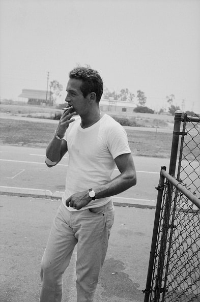 A man demonstrates how to wear a white t-shirt with style, pairing it with light pants as he stands near a chain-link fence, smoking a cigarette, with a road and buildings in the background.