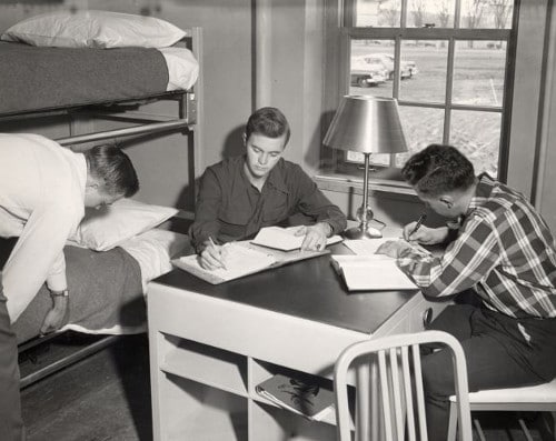 Three Vintage college students at desk in dorm room two are studying while third one is setting his bed.