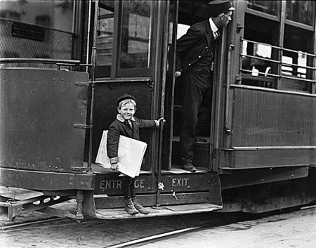 A kid is standing on the door of a trolley car.