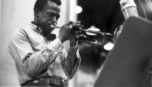 A black and white photo of a man playing a trumpet in a tucked-in shirt.