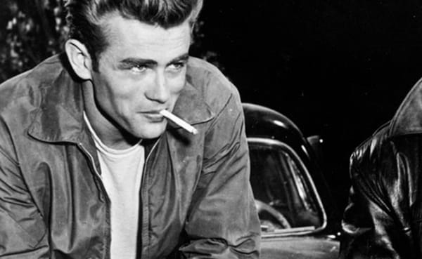 A young man with effortless style leans forward, wearing a white t-shirt and jacket, a cigarette in his mouth; a car is visible in the background.