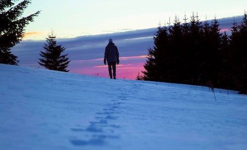 Person walking on a snow-covered slope at dusk with a colorful sky in the background, inspired by "Befriending Winter" from Podcast #856.
