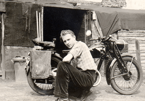 A man sitting on a motorcycle, his first motorcycle, in front of a shack.